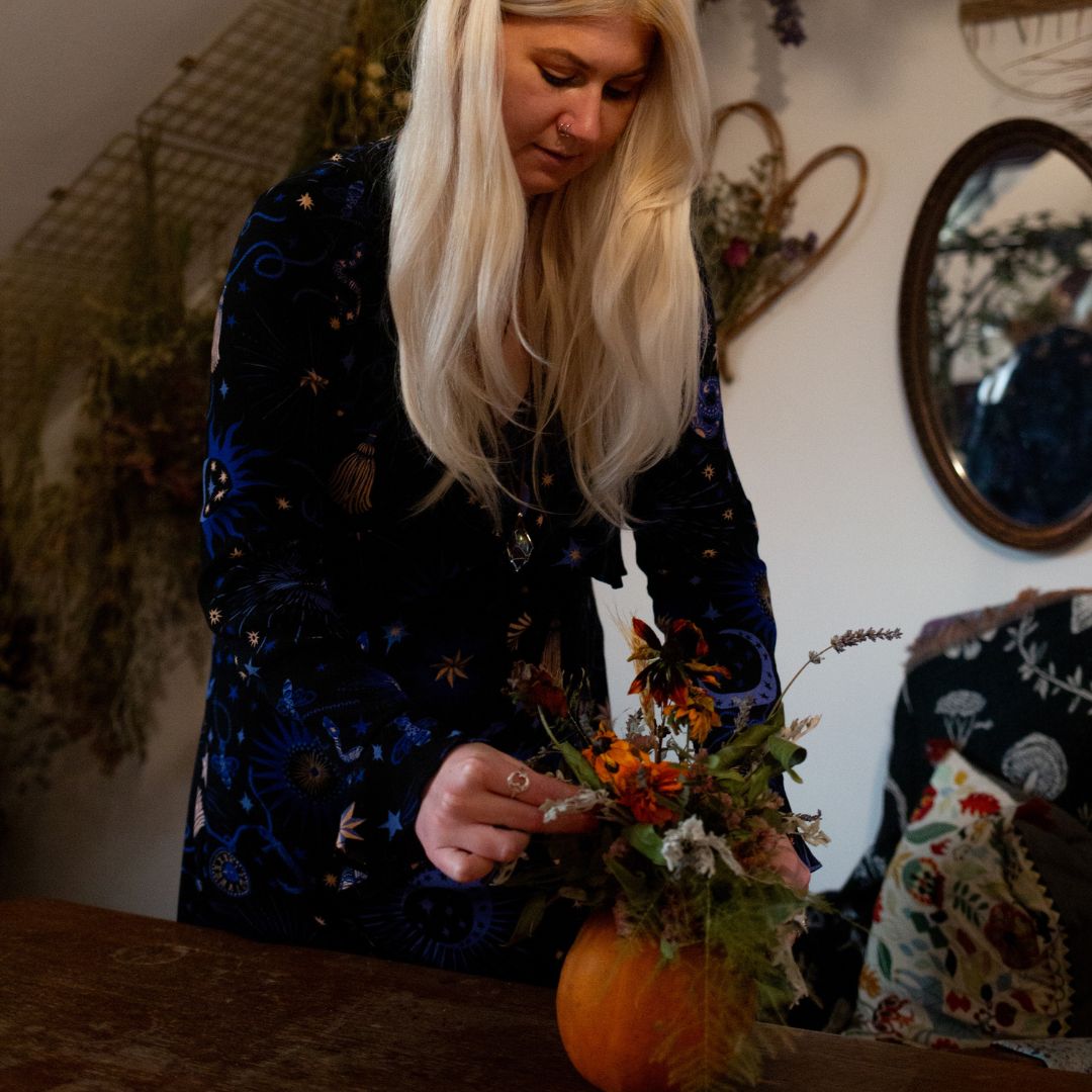 Thistle and Moonlace flower studio, broomstick, mirror, dried florals hung from the ceiling and walls surrounding a blonde haired woman making a floral arrangement on a pumpkin.