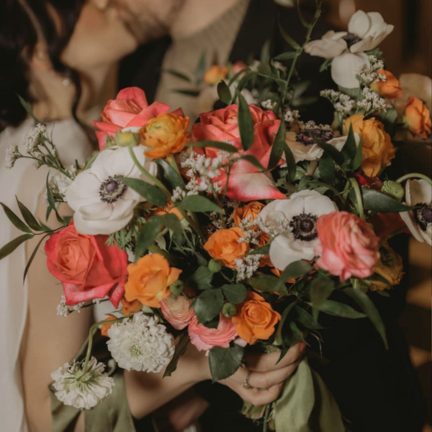 Wedding couple kissing with their fresh floral bouquet in the main focus. White anemones, pink and orange  roses, white scabiosa, soft pink ranunculus and white limonium.