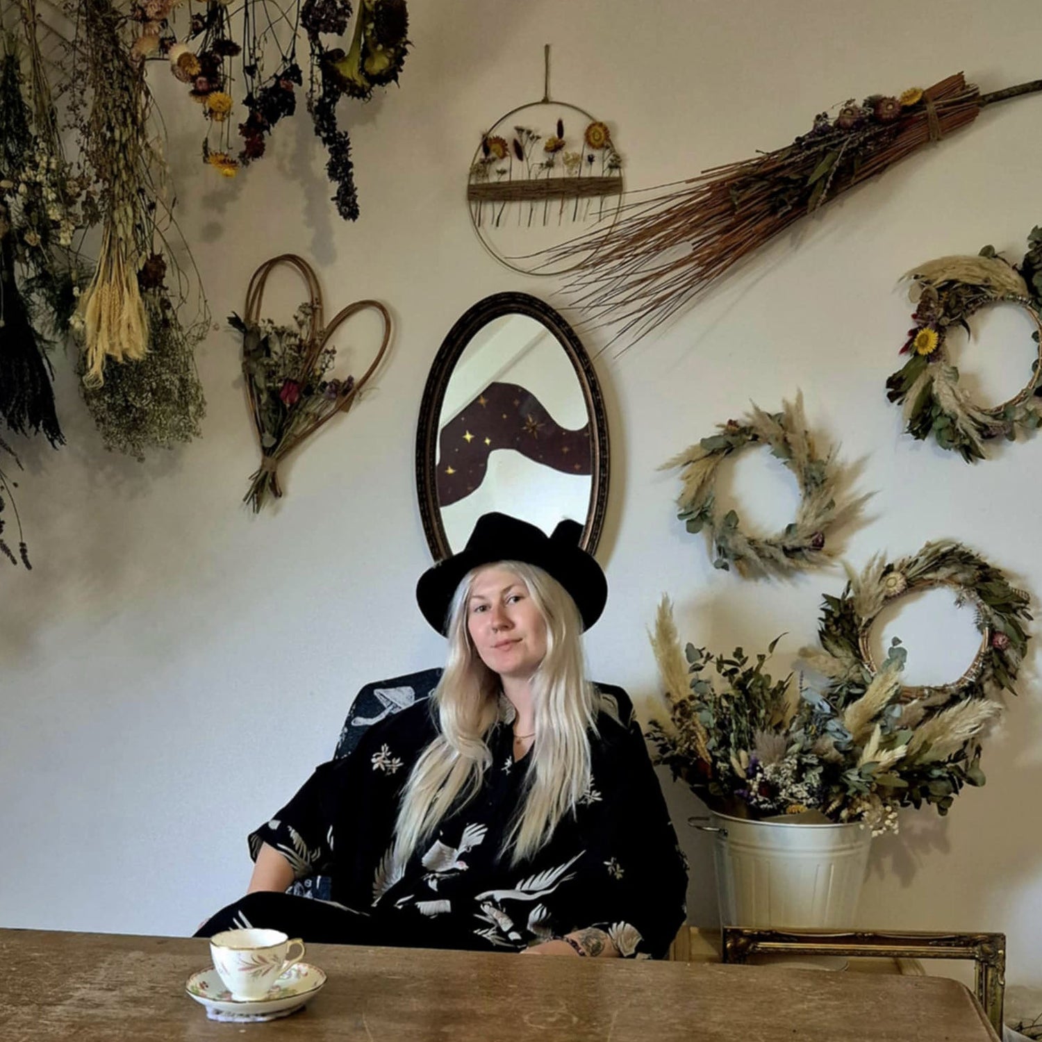 Woman in a black hat sitting at a table with decorative and enchanting floral  elements on the wall behind her.