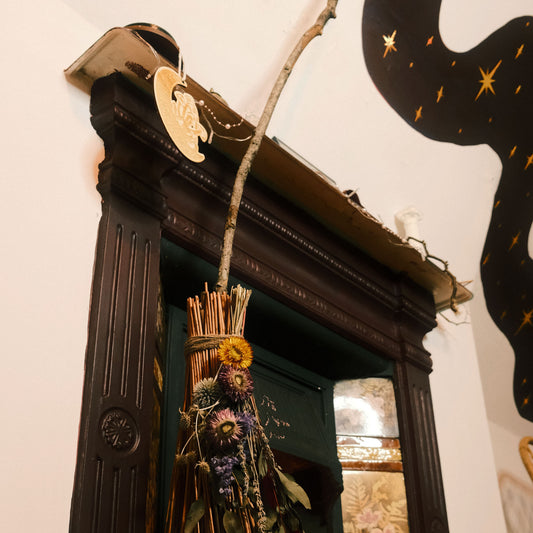 Decorative broom with flowers hanging leaned against an Edwardian fireplace in a dark academia room with a purple moody paint and gold stars on the wall.