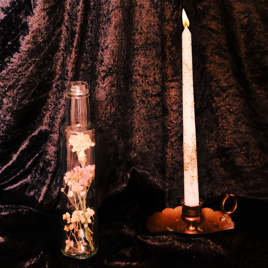 Glass bottle with dried flowers and lit candle against a dark curtain background