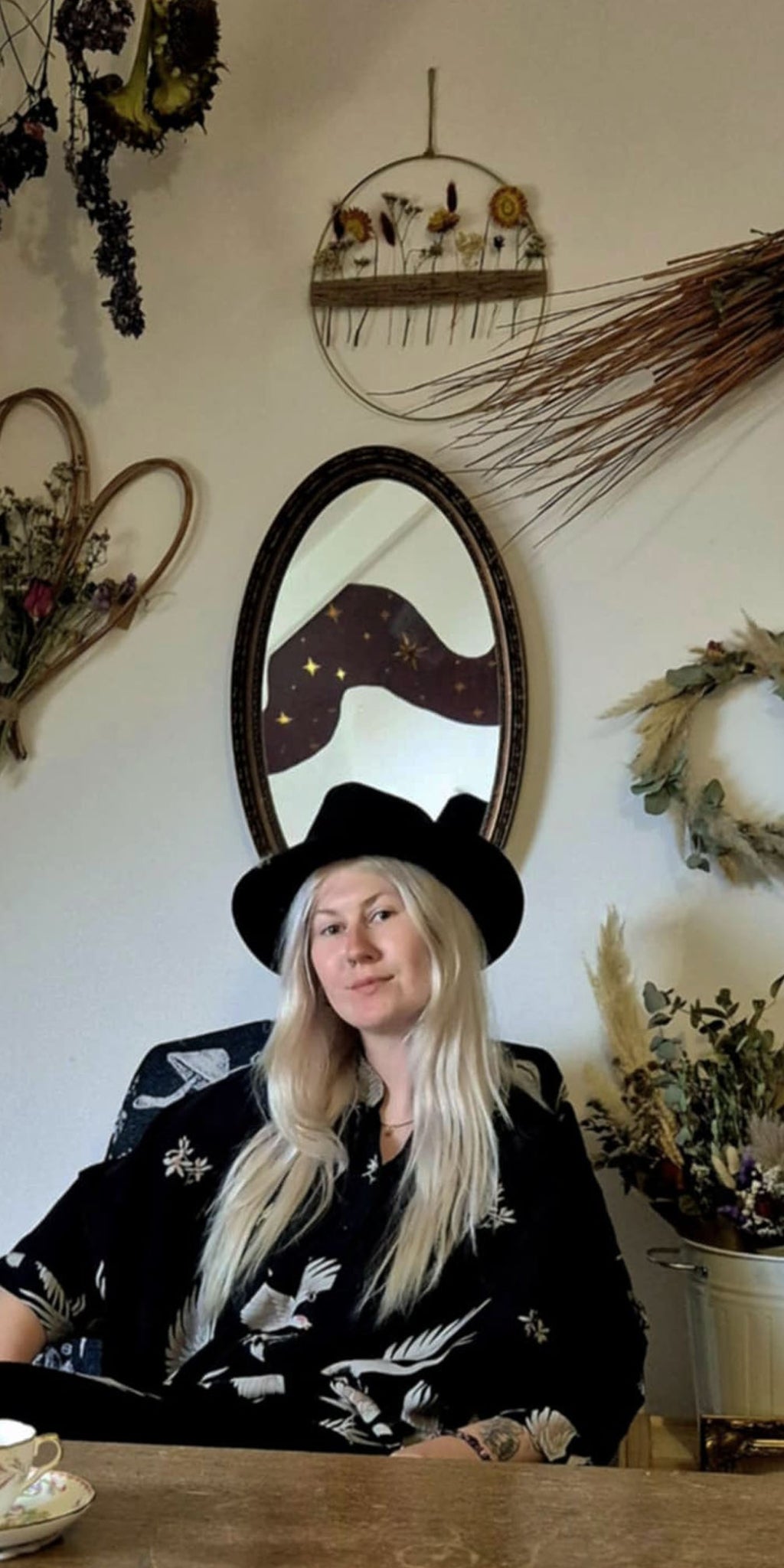 Woman in a black hat sitting at a table with decorative and enchanting floral  elements on the wall behind her.